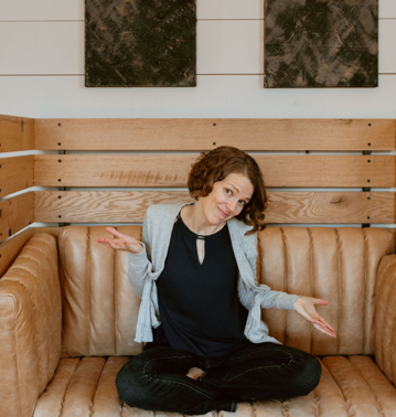 Lisa McH, Digital Organizer and creator of the Tame Your Inbox Self-Paced Course, sits on a carmel colored leather couch with wooden slats wearing in a gray sweater and black shirt.